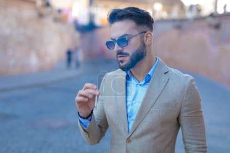 Téléchargez les photos : Portrait of elegant young businessman with sunglasses looking to side and smoking outdoor in an old city from Transylvania - en image libre de droit