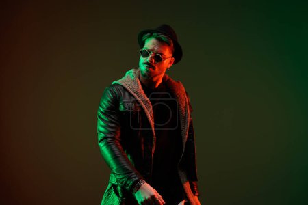 Téléchargez les photos : Portrait de jeune homme décontracté regardant loin et posant avec un style cool dans un jeu de lumières colorées - en image libre de droit