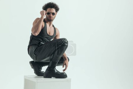 Téléchargez les photos : Sexy casual homme accroupi sur la chaise et des lunettes de fixation, portant un costume en cuir dans une pose de mode - en image libre de droit