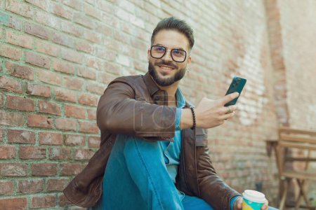 Téléchargez les photos : Portrait de jeune homme décontracté s'amusant à texter et boire, portant des lunettes et une veste en cuir, en plein air dans une vieille ville médiévale - en image libre de droit