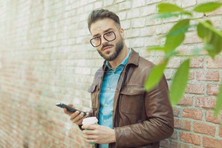 Téléchargez les photos : Portrait d'un homme décontracté attrayant textant au téléphone et tenant une boisson contre un mur, portant des lunettes et une veste en cuir, en plein air dans une vieille ville médiévale - en image libre de droit