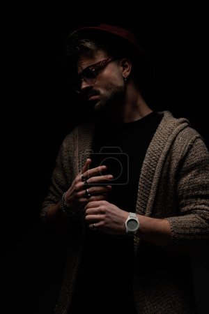 Téléchargez les photos : Portrait de jeune homme décontracté posant avec une ambiance difficile, portant un chapeau bordeaux, des lunettes et un manteau de laine en arrière-plan studio sombre - en image libre de droit