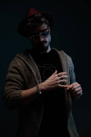 Foto de Retrato de chico casual guapo con estilo agradable sosteniendo su mano, usando un sombrero de color burdeos, gafas y abrigo de lana en el fondo del estudio oscuro - Imagen libre de derechos