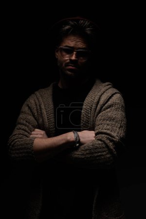Photo for Portrait of attractive casual model crossing his arms at chest, wearing a burgundy hat, eyeglasses and wool coat in dark studio background - Royalty Free Image