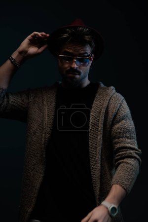 Photo for Portrait of handsome fashion man arranging his burgundy hat, wearing a burgundy hat, eyeglasses and wool coat in dark studio background - Royalty Free Image