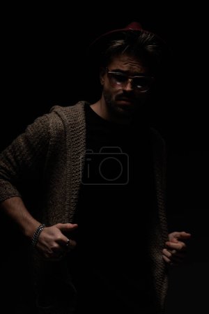 Foto de Retrato de un sexy hombre casual arreglando su chaqueta, usando un sombrero de color burdeos, anteojos y abrigo de lana en el fondo oscuro del estudio - Imagen libre de derechos