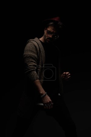 Foto de Retrato de chico casual guapo posando en una postura sexy, usando un sombrero de color burdeos, gafas y abrigo de lana en el fondo del estudio oscuro - Imagen libre de derechos