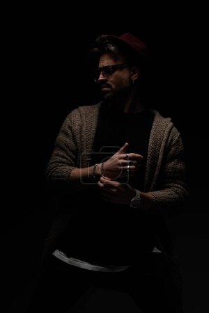 Photo for Portrait of attractive casual model arranging his bracelet and looking away, wearing a burgundy hat, eyeglasses and wool coat in dark studio background - Royalty Free Image