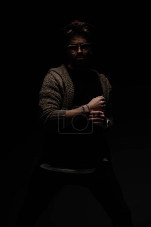 Téléchargez les photos : Portrait de modèle masculin sexy dans une position difficile fixant son bracelet, portant un chapeau bordeaux, lunettes et manteau de laine en arrière-plan studio sombre - en image libre de droit