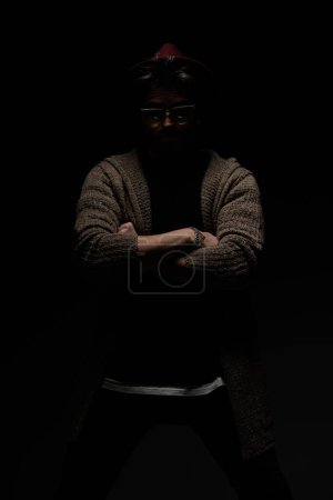 Foto de Retrato de un joven tipo casual cruzando los brazos como un macho, con un sombrero de color burdeos, anteojos y abrigo de lana en el fondo oscuro del estudio - Imagen libre de derechos