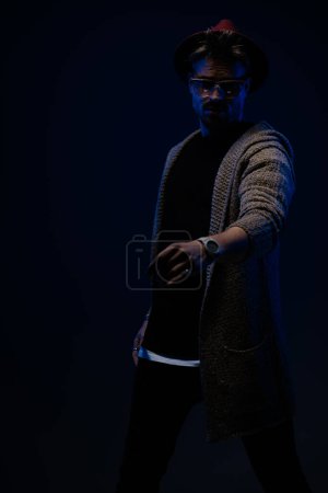 Foto de Retrato de modelo masculino atractivo posando con los brazos sueltos alrededor del cuerpo, usando un sombrero de color burdeos, gafas y abrigo de lana en el fondo oscuro del estudio - Imagen libre de derechos