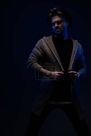Foto de Retrato del guapo tipo de la moda tirando lentamente de su chaqueta, usando un sombrero de color burdeos, gafas y abrigo de lana en el fondo oscuro del estudio - Imagen libre de derechos