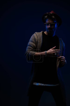Téléchargez les photos : Portrait de mec décontracté sexy posant dans une position dramatique, portant un chapeau bordeaux, des lunettes et un manteau de laine en arrière-plan studio sombre - en image libre de droit