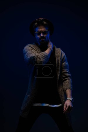 Foto de Retrato del joven hombre de la moda frotándose lentamente el cuello, usando un sombrero de color burdeos, gafas y abrigo de lana en el fondo oscuro del estudio - Imagen libre de derechos