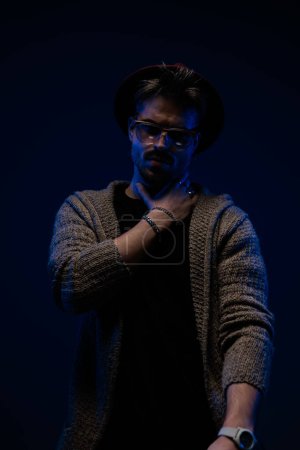Foto de Retrato de un hombre casual guapo tocándose el cuello con un aspecto penetrante, con un sombrero de color burdeos, anteojos y abrigo de lana en el fondo oscuro del estudio - Imagen libre de derechos