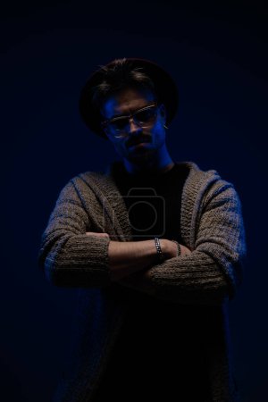Foto de Retrato de un atractivo tipo casual con poderosa presencia cruzando los brazos, con un sombrero de color burdeos, gafas y abrigo de lana en el fondo oscuro del estudio - Imagen libre de derechos