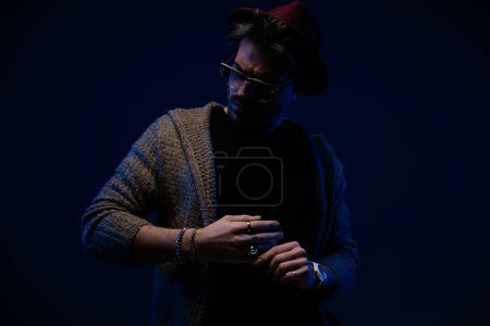 Foto de Retrato de un joven modelo masculino con un ambiente dramático que fija su anillo, con un sombrero de color burdeos, anteojos y abrigo de lana en el fondo oscuro del estudio - Imagen libre de derechos