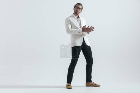 Téléchargez les photos : Sexy mode gars portant veste blanche costume frotter les paumes et debout en face de fond gris - en image libre de droit
