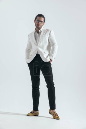 Foto de Hombre joven de moda con traje de chaqueta blanca cogido de la mano en los bolsillos y de pie delante del fondo gris en el estudio - Imagen libre de derechos