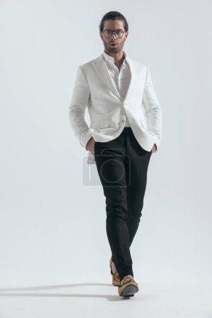 Téléchargez les photos : Homme de mode sexy avec des lunettes tenant la main dans les poches et marchant devant fond gris - en image libre de droit