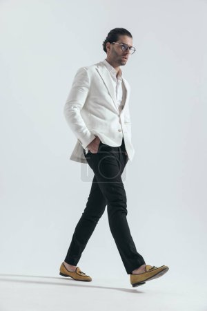 Photo for Side view of cool fashion man with glasses walking with hands in pockets and looking to side on grey background in studio - Royalty Free Image