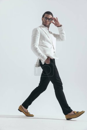 Téléchargez les photos : Vue latérale de l'homme de mode sexy marchant avec la main dans les poches, tenant la main au front et posant sur fond gris - en image libre de droit