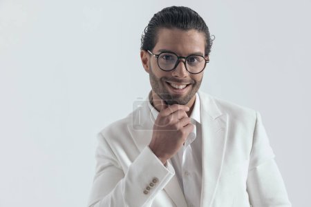 Téléchargez les photos : Portrait d'un beau jeune homme en costume blanc touchant le menton, réfléchissant et souriant sur fond gris - en image libre de droit