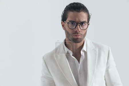 Téléchargez les photos : Homme de mode cool avec des lunettes portant un costume de veste blanche et chemise à col ouvert devant fond gris - en image libre de droit