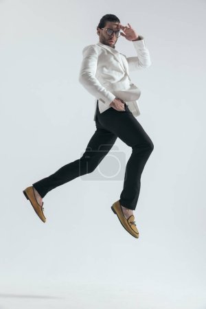 Photo for Cool young man with glasses holding hand in pocket and touching forehead while jumping in the air on grey background - Royalty Free Image