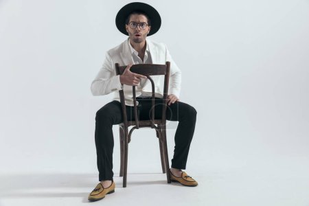 Téléchargez les photos : Jeune homme sexy avec chapeau et lunettes faisant un visage choqué tout en étant assis sur une chaise en bois devant fond gris - en image libre de droit