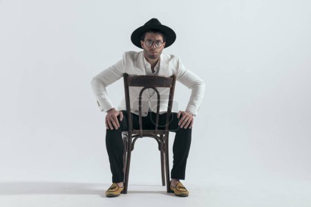 Téléchargez les photos : Homme de mode sexy avec chapeau assis sur une chaise en bois avec paumes sur les genoux en face de fond gris - en image libre de droit