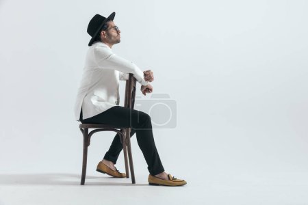 Téléchargez les photos : Vue latérale du gars cool de la mode avec chapeau et lunettes de soleil assis sur une chaise en bois et regardant vers le côté en face de fond gris en studio - en image libre de droit