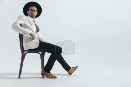 Téléchargez les photos : Cool homme de mode avec chapeau et lunettes tenant le bras sur la chaise et assis devant fond gris - en image libre de droit