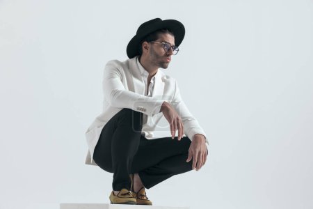 Photo for Cool fashion guy with hat and glasses holding arms on knees and crouching in front of grey background in studio - Royalty Free Image