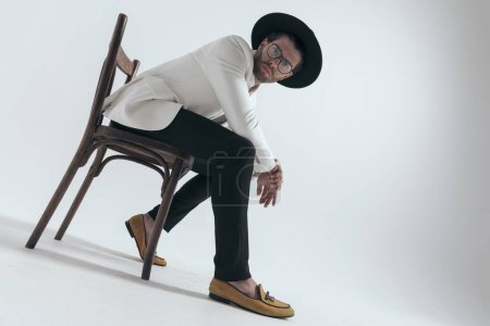 Photo for Bottom view of cool young guy with hat and glasses holding elbows on knees and sitting on grey background - Royalty Free Image