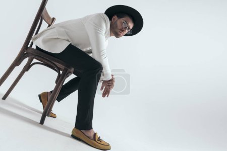 Téléchargez les photos : Vue du bas du jeune homme sexy tenant les coudes sur les genoux et assis sur une chaise en bois devant un fond gris en studio - en image libre de droit