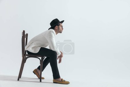 Téléchargez les photos : Vue latérale de la mode jeune modèle avec chapeau et lunettes regardant vers le côté, tenant les coudes sur les genoux et assis sur une chaise en bois sur fond gris - en image libre de droit