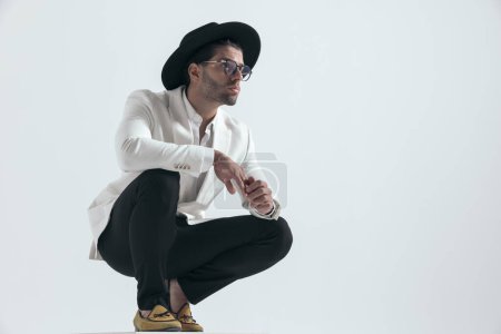 Photo for Sexy attractive young man with hat and glasses crouching and looking to side while touching fingers and holding arms on knees on grey background - Royalty Free Image