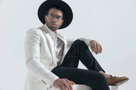 Téléchargez les photos : Cool homme de mode avec chapeau et lunettes assis et tenant le coude sur le genou devant fond gris - en image libre de droit