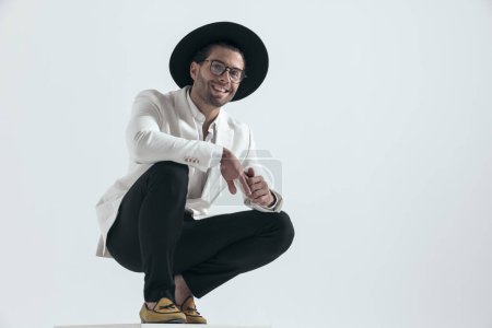 Photo for Happy elegant man with hat and glasses crouching and smiling, holding arms on knees and touching fingers on grey background - Royalty Free Image