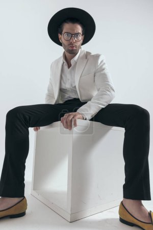 Téléchargez les photos : Homme de mode sexy portant chapeau et lunettes et posant d'une manière cool tout en étant assis sur fond blanc - en image libre de droit