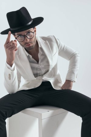 Téléchargez les photos : Cool mode sexy homme avec des lunettes en veste blanche costume ajustement chapeau tout en étant assis en face de fond gris - en image libre de droit