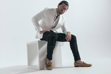 Photo for Attractive man with glasses in white jacket suit with open collar shirt sitting and holding arms on thigh in front of grey background - Royalty Free Image
