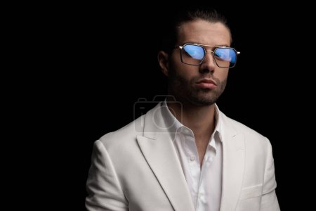 Téléchargez les photos : Beau gars élégant avec chemise à col ouvert et lunettes portant costume blanc posant en face de fond noir - en image libre de droit
