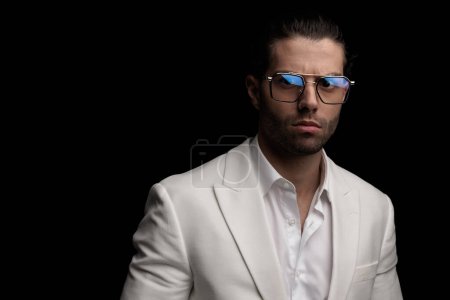 Téléchargez les photos : Élégant jeune homme avec des lunettes portant chemise et veste col ouvert blanc et posant de manière confiante sur fond noir - en image libre de droit