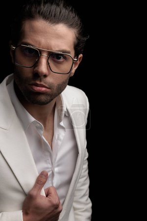Téléchargez les photos : Découpez l'image de l'homme de mode sexy avec des lunettes tenant costume veste blanche et posant devant fond noir - en image libre de droit