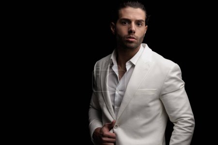 Téléchargez les photos : Jeune homme de mode arrangeant costume blanc et posant d'une manière confiante devant fond noir - en image libre de droit