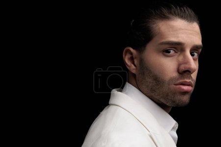 Side view of sexy young man in white suit with open collar shirt posing in front of black background, close up