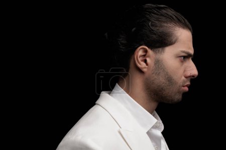 Téléchargez les photos : Jeune homme sexy avec des cheveux bouclés portant un costume blanc et regardant vers le côté tout en posant sur fond noir, photo rapprochée - en image libre de droit
