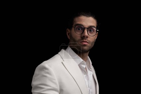 Téléchargez les photos : Vue latérale d'attrayant jeune homme avec des lunettes en costume blanc étant surpris en face de fond noir - en image libre de droit